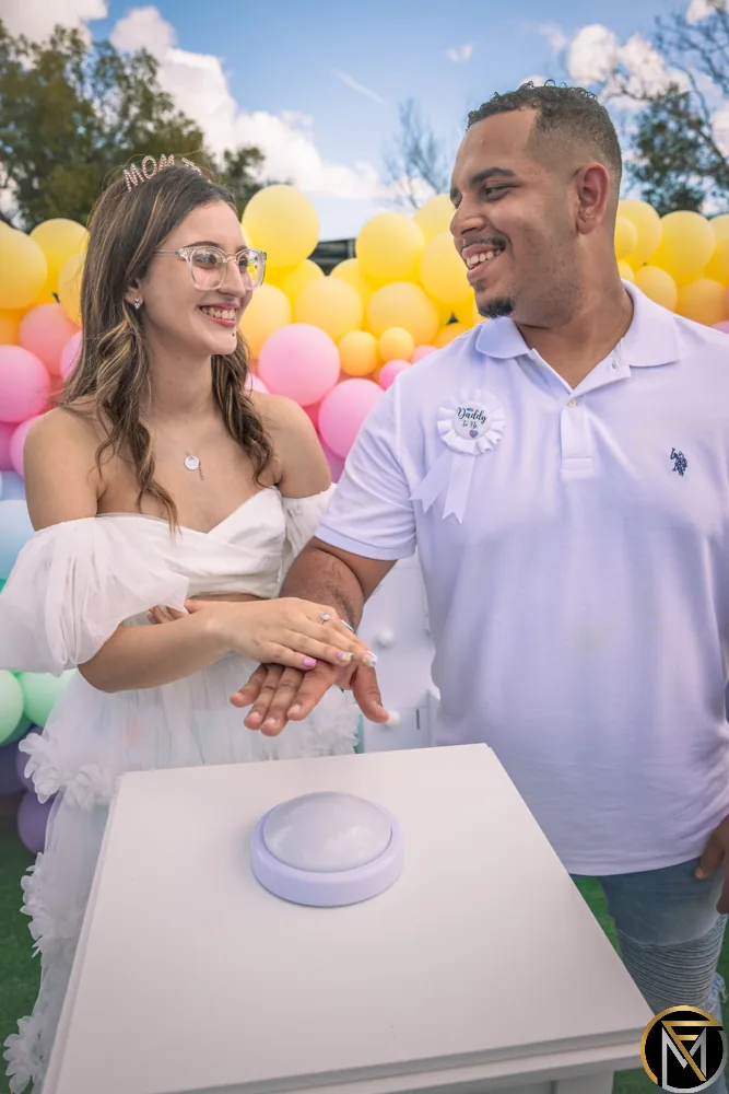 A couple smiles at each other with their hands on the button at their gender reveal party