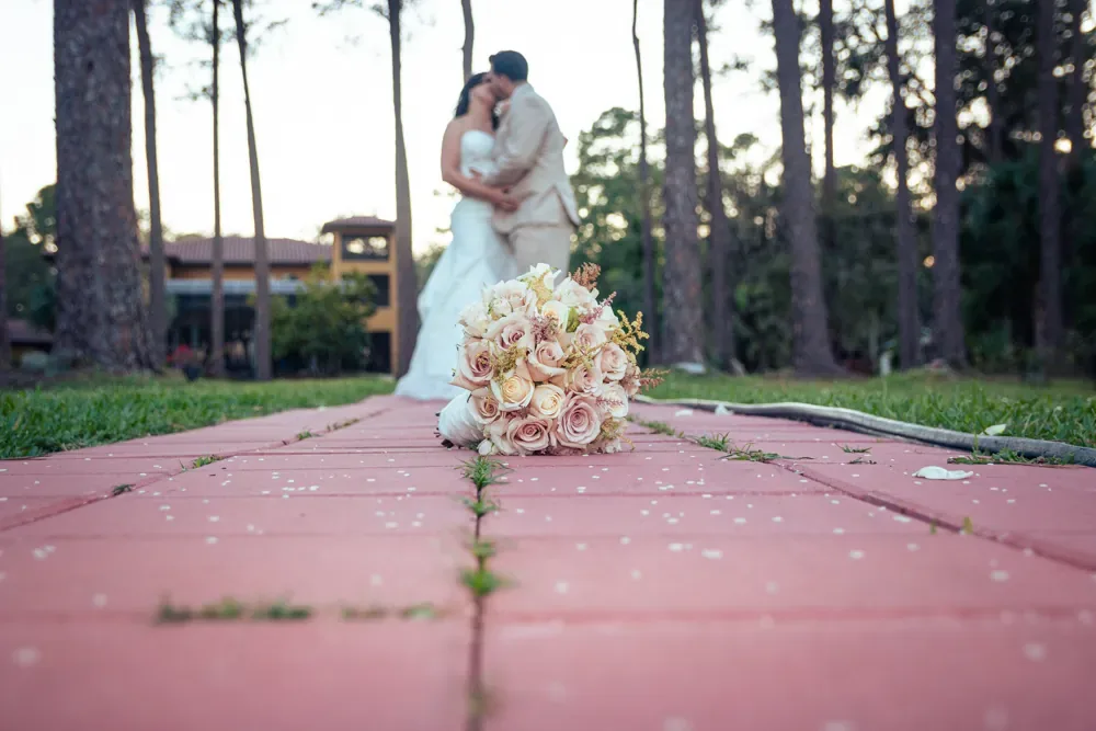 A bride and groom share a kiss under tall pines, with her bouquet of blush roses resting on the brick path in front of them