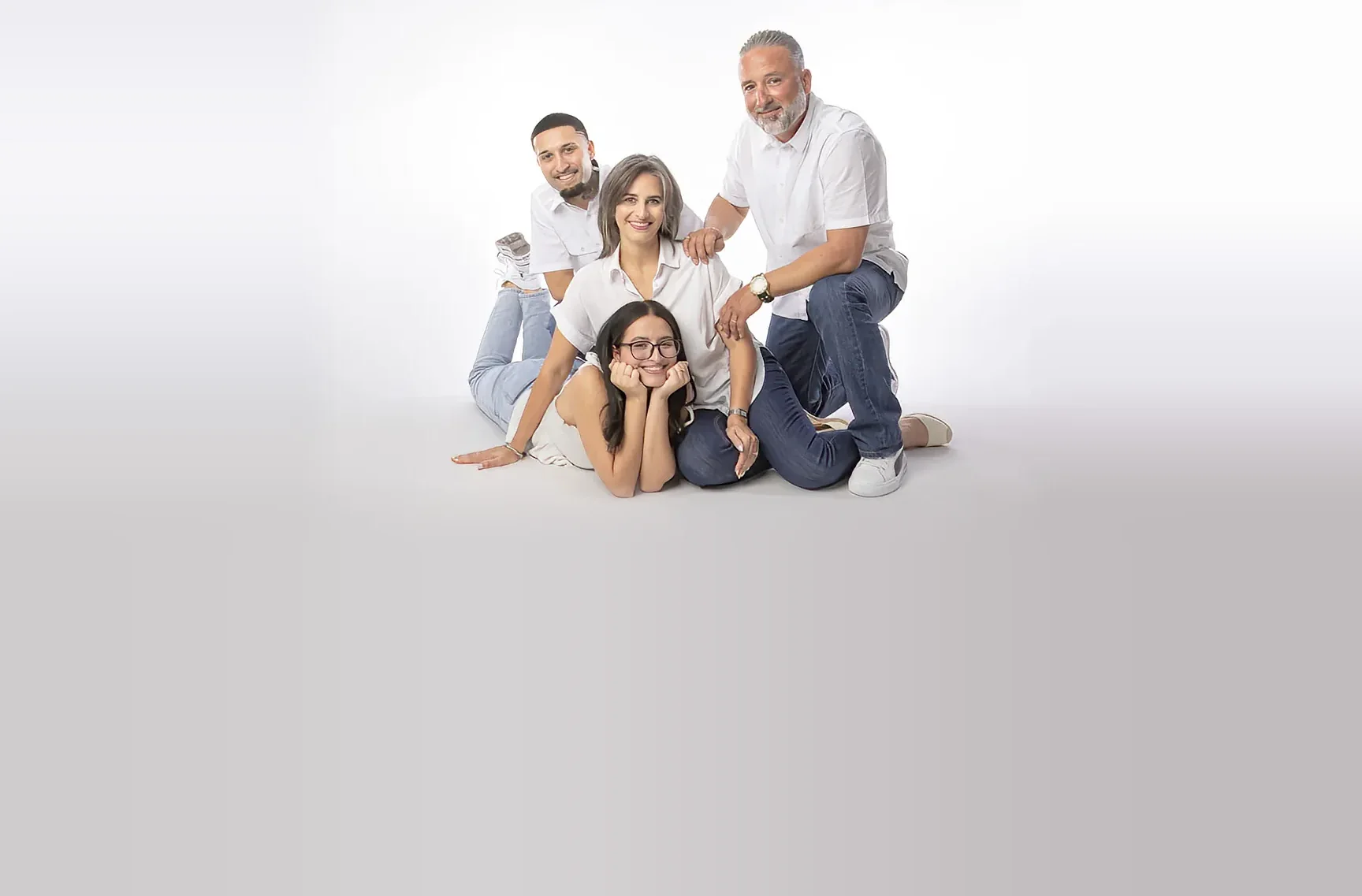 A family of four in white shirts and denim poses together, smiling, on a white studio floor