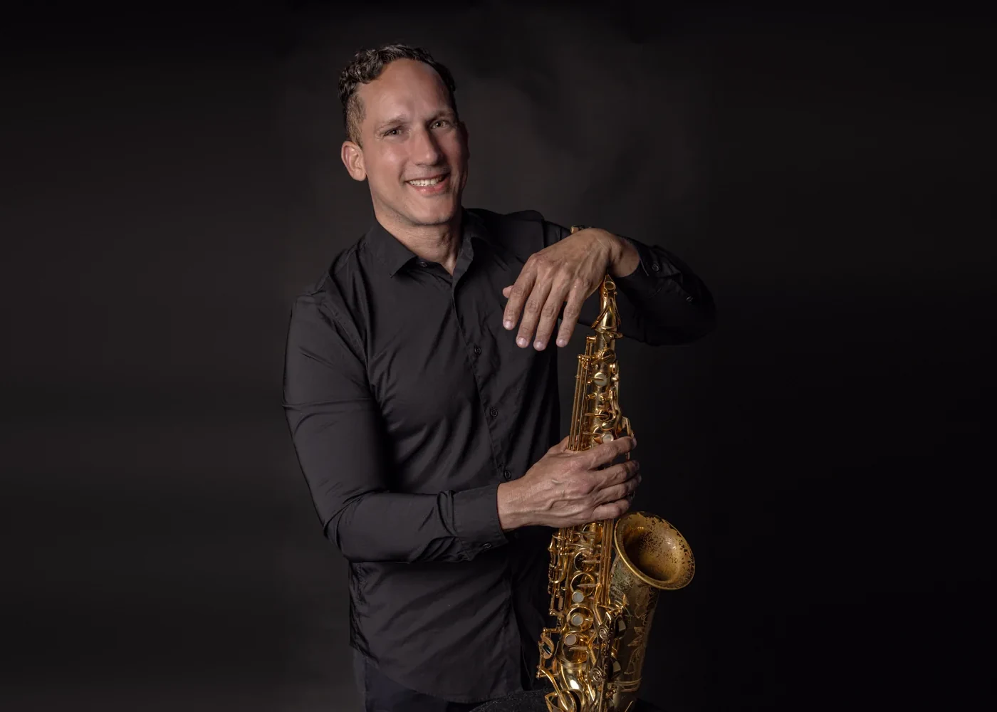 A musician in a black shirt smiles while leaning on his saxophone in a dark studio portrait